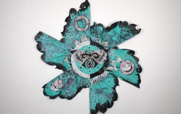 An abstract wall sculpture featuring a vibrant turquoise design with intricate patterns and circular elements. The piece resembles a flower or starburst, combining organic shapes and textures, creating a dynamic visual impact against a plain background.