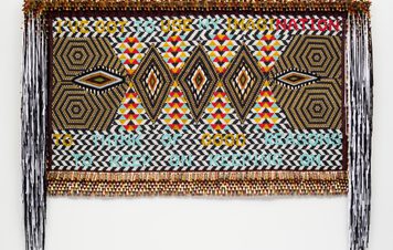 A vibrant textile artwork featuring geometric patterns in bold colours, including red, yellow, and black. The piece includes the text "I've got one imagination to good reason" woven into the design, framed with decorative fringes.