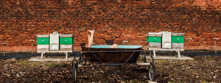 A leg sticking out of a bath tub on wheels in front of a brick wall outdoors between two bee hives.