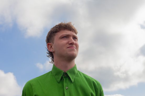 A person with short blonde hair wearing a green shirt in front of a cloudy sky.