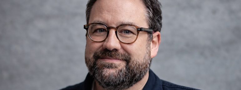 A white man with short brown hair, short beard and glasses looks directly into the camera.