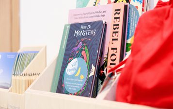 A close-up of a bookshelf displaying various books. Prominent titles include "Here Be Monsters" and "Rebel Folk." The shelf is made of light wood, and there are colourful bags and other items in the background, creating a vibrant, inviting atmosphere.