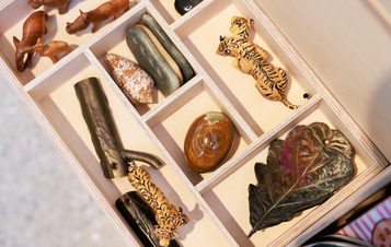 A wooden box divided into compartments, containing various small objects: carved animals, smooth stones, a shiny shell, a wooden stick, a decorative leaf, and two tiger figurines, all arranged neatly within the sections.