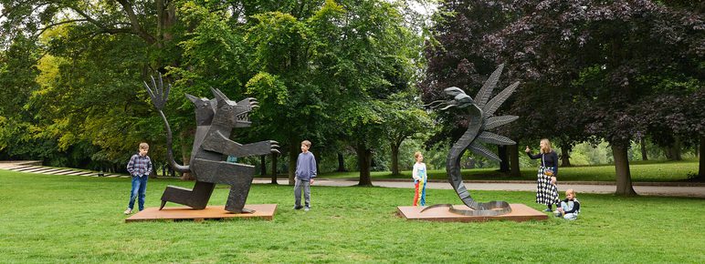 Two large abstract sculptures stand in a green park, one resembling a bear with outstretched arms and the other a three headed snake. Four children play nearby, exploring the art amidst lush trees and a serene landscape.