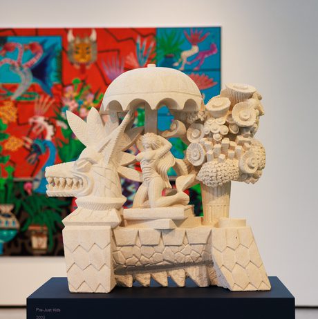 A stone sculpture depicting a mythical creature with a canopy, featuring a figure and stylised trees. In the background, a vibrant, abstract painting with bold colours and floral motifs adds contrast to the artwork.