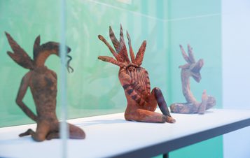 Three intricately carved sculptures are displayed on a white shelf within a glass case. Each figure features distinct features, such as feathered headdresses and unique postures, set against a softly coloured background.