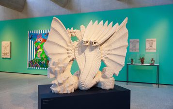 A large, white sculptural piece resembling a mythical creature with expansive wings is displayed in an art gallery. The background features vibrant green walls, with colourful artworks and framed sketches visible on the walls.