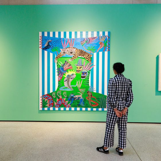 A person in a black and white checked outfit stands in front of a vibrant artwork featuring bold colours and patterns, flanked by two white sculptures on a bright green wall in an art gallery.