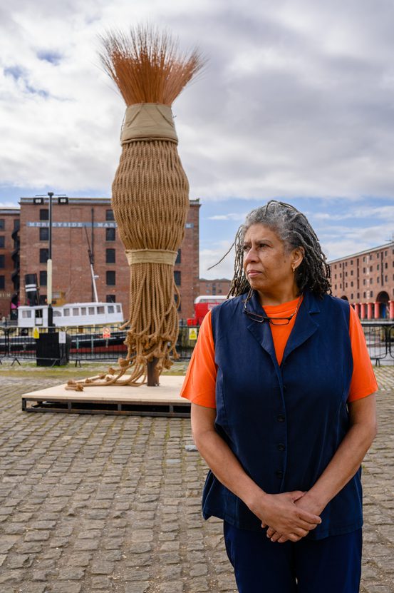 A woman with long, grey hair stands in front of a large woven sculpture resembling a broom. She wears an orange top and navy vest, with a cobbled waterfront and historic buildings in the background under a cloudy sky.