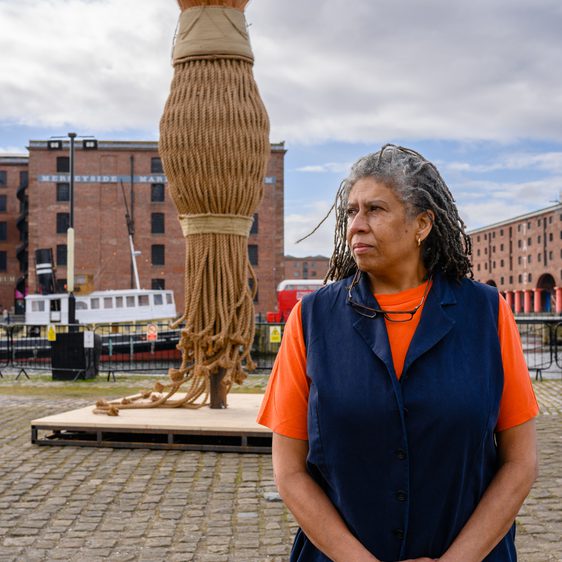 A woman with long, grey hair stands in front of a large woven sculpture resembling a broom. She wears an orange top and navy vest, with a cobbled waterfront and historic buildings in the background under a cloudy sky.