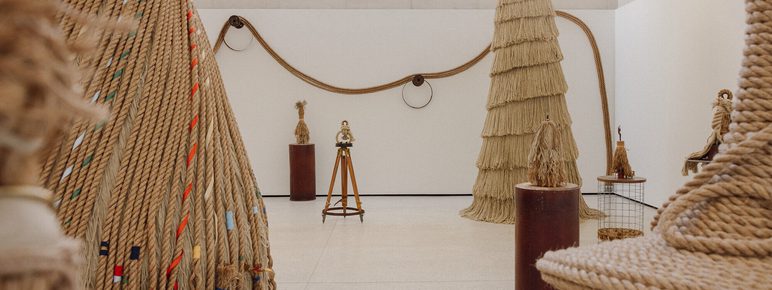 An art gallery featuring large sculptures made of rope. The space is bright and minimalistic, with various rope structures of different heights and textures. A wooden tripod stands in the centre, surrounded by intricate rope designs.