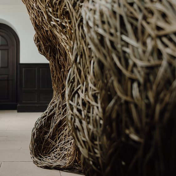 A close-up view of a large, woven sculpture made of natural materials, positioned in a spacious interior. In the background, a dark wooden door with an arched top is visible, framed by light-coloured walls and flooring.