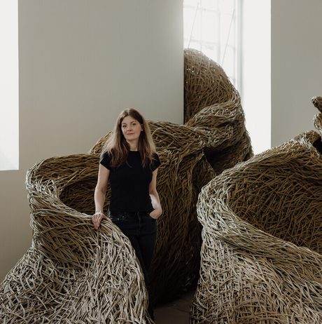 A woman stands beside large, intricately woven sculptures made of natural materials. The sculptures resemble organic forms and are set against a bright, minimalist gallery space with large windows. The woman wears a black top and jeans.