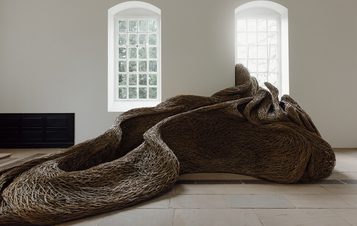 A large, organic sculpture made of intertwined natural materials sits in a bright, minimalist gallery. The piece resembles flowing, textured forms and is illuminated by natural light streaming through tall windows.