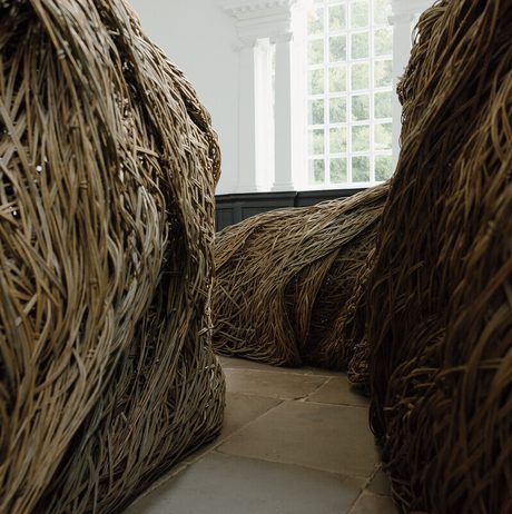 An interior space featuring large, intricately woven structures made of natural materials. The formations create winding pathways, with light filtering through a large window, highlighting the textures and organic shapes of the artwork.