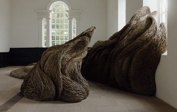 A large, organic sculpture made of intertwined natural materials resembles flowing waves or abstract forms. It is positioned in a bright room with large windows, contrasting with the stone floor.