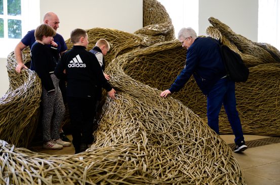 A group of four people, including two children, interact with a large, woven sculpture made of natural materials. One adult observes while another touches the textured surface. The setting appears to be a bright, spacious gallery.