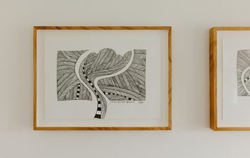 A framed black and white artwork featuring a stylised tree with intricate leaf patterns and winding lines. The piece is displayed on a light-coloured wall, alongside another similar frame, showcasing a minimalist and artistic aesthetic.
