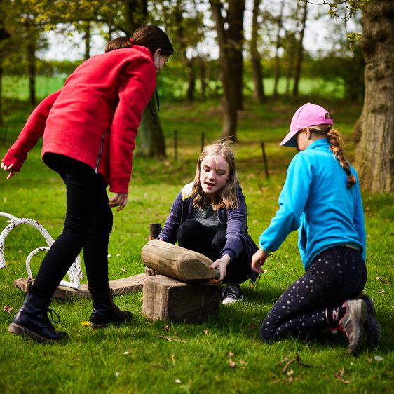 Three children building wooden log sculptures.
