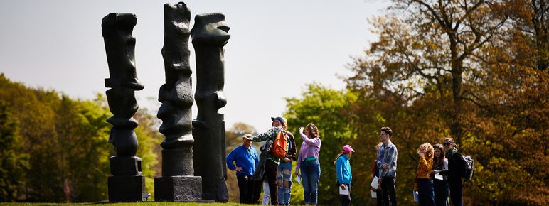 A large group of people on a tour, admiring a Henry Moore sculpture.