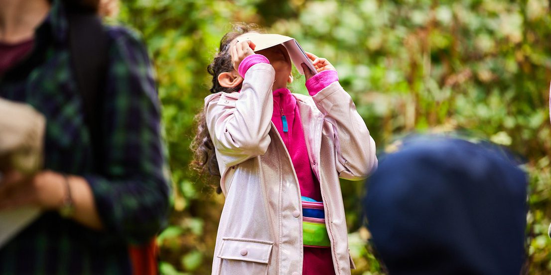 A child wearing a pink rain coat, looking through a paper viewfinder outdoors
