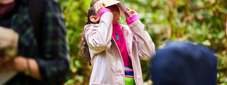 A child wearing a pink rain coat, looking through a paper viewfinder outdoors