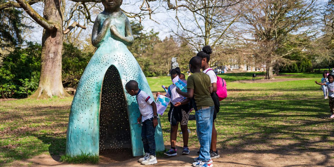 A group of children explore a large, whimsical sculpture of a woman in a dress, set in a park. The sculpture has a textured surface and is surrounded by trees. The children are engaged, with one examining the sculpture closely while others hold maps.