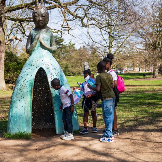A group of children explore a large, whimsical sculpture of a woman in a dress, set in a park. The sculpture has a textured surface and is surrounded by trees. The children are engaged, with one examining the sculpture closely while others hold maps.