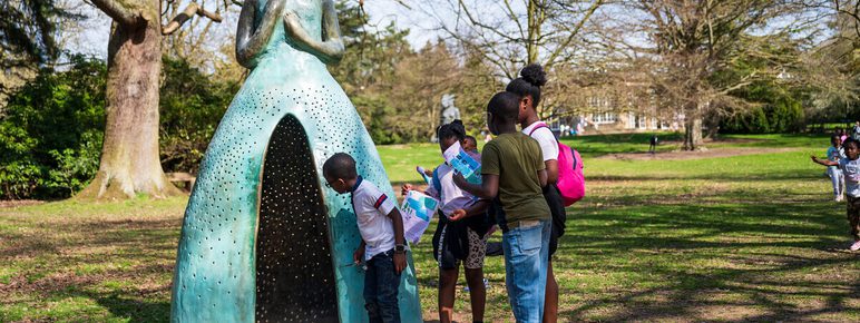 A group of children explore a large, whimsical sculpture of a woman in a dress, set in a park. The sculpture has a textured surface and is surrounded by trees. The children are engaged, with one examining the sculpture closely while others hold maps.