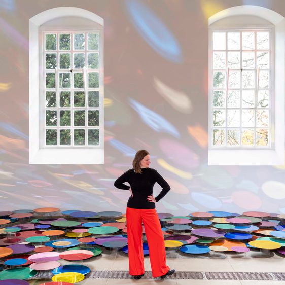 A woman wearing a black top and red trousers standing inside an installation of colourful mirrored discs displayed at floor level. Large windows and colourful reflections are on the wall behind her.