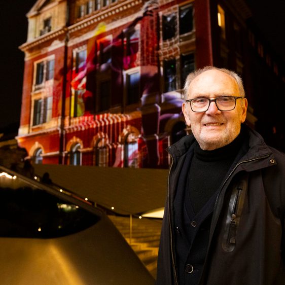 A man with grey hair and glasses, standing in front of a building which has a digital artwork projected on to it.
