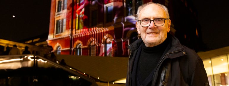 A man with grey hair and glasses, standing in front of a building which has a digital artwork projected on to it.