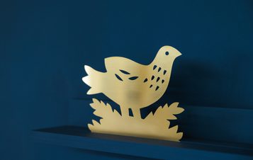 A brass cut out of a bird displayed on a shelf against a blue wall.