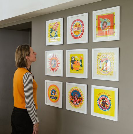A woman wearing an orange vest looks at a group of colourful frames artworks displayed in a grid on a grey wall.