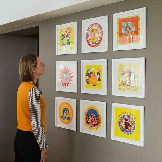 A woman wearing an orange vest looks at a group of colourful frames artworks displayed in a grid on a grey wall.