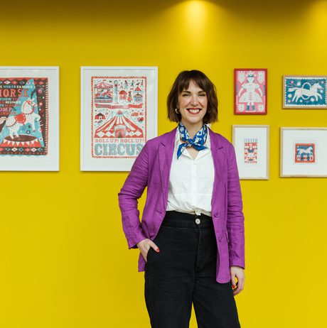 A smiling woman with short brown hair stands in front of a display of colourful framed artworks on a bright yellow wall.