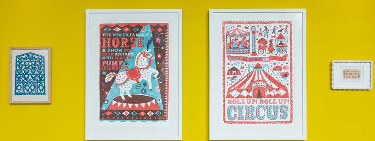 A display of colourful framed artworks on a bright yellow wall.