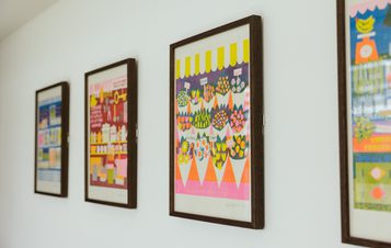 A row of colourful prints in dark wood frames, displayed on a white wall.