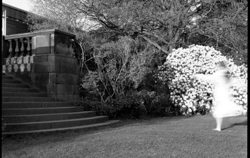 A black and white image of a person wearing a white dress running towards a set of steps in a garden.