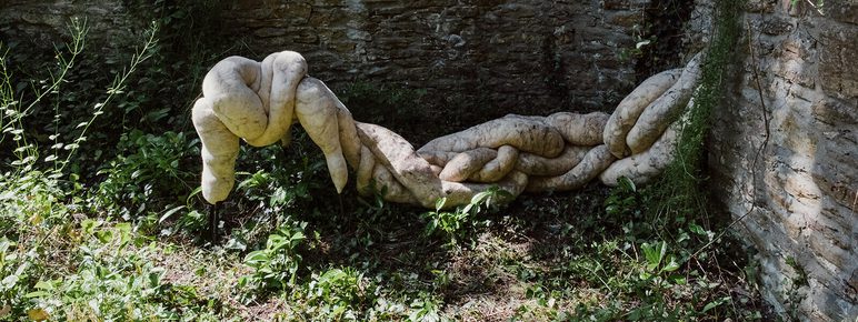 A surreal sculpture resembling intertwined, organic forms lies against a stone wall, surrounded by lush greenery. The soft, pale textures contrast with the rugged stone, creating an intriguing visual interplay between nature and art.
