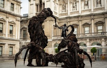 A large, abstract sculpture made of intertwined dark fabric forms two towering figures that appear to reach towards a central statue of a man. The setting is an elegant historic building, with classical architecture in the background.