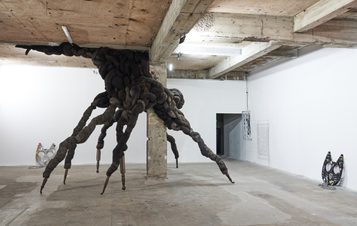 An abstract sculpture resembling a large, twisted creature with multiple limbs emerges from a concrete pillar in a minimalist gallery space. The floor is bare, and the walls are white, enhancing the striking presence of the artwork.