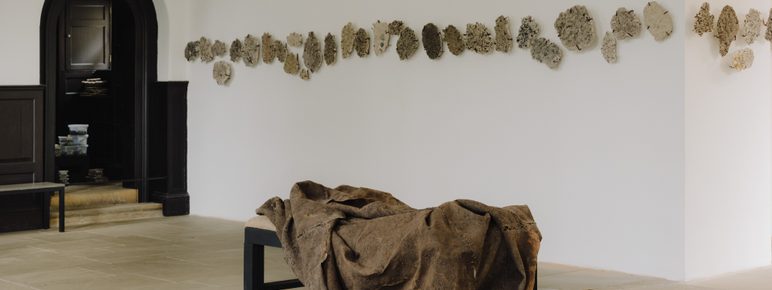 A minimalist art gallery featuring a textured, draped sculpture on a black bench. The wall behind displays a series of abstract, organic shapes in muted tones, creating a contrast with the clean, white space and dark architectural elements.