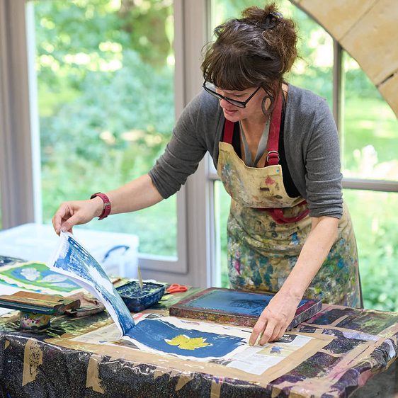 A woman wearing a paint covered apron, demonstrates a printing technique using leaves in a brightly lit studio with a window behind her.