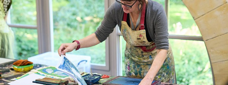 A woman wearing a paint covered apron, demonstrates a printing technique using leaves in a brightly lit studio with a window behind her.