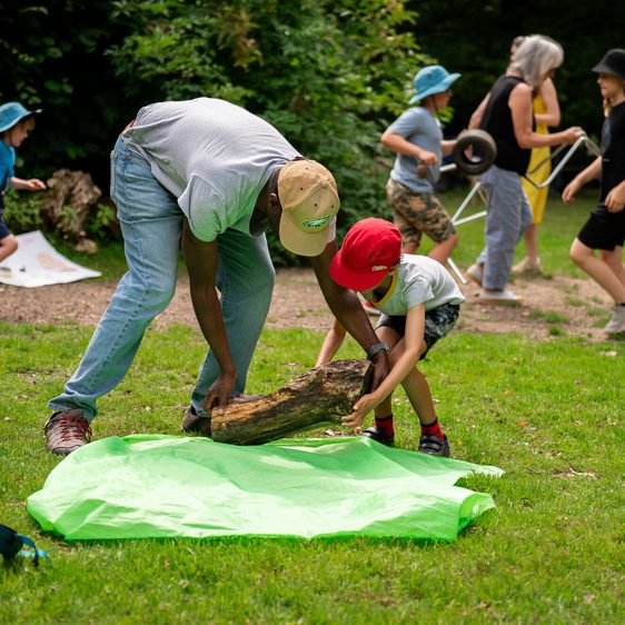 A man and a young boy lift a large piece of wood on to a green tarp in a park. In the background there are several other children and adults collecting objects for sculpture building.
