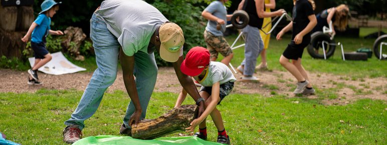 A man and a young boy lift a large piece of wood on to a green tarp in a park. In the background there are several other children and adults collecting objects for sculpture building.