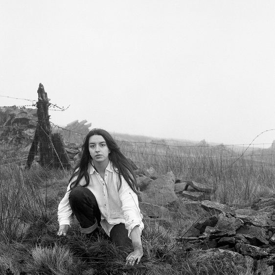 A woman wearing a white shirt crouches in a field of long grasses, with wooden structures in the distance.