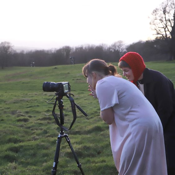 Two people, one wearing a red woolly hat, the other a nightdress crouched over looking into the lense of a camera.