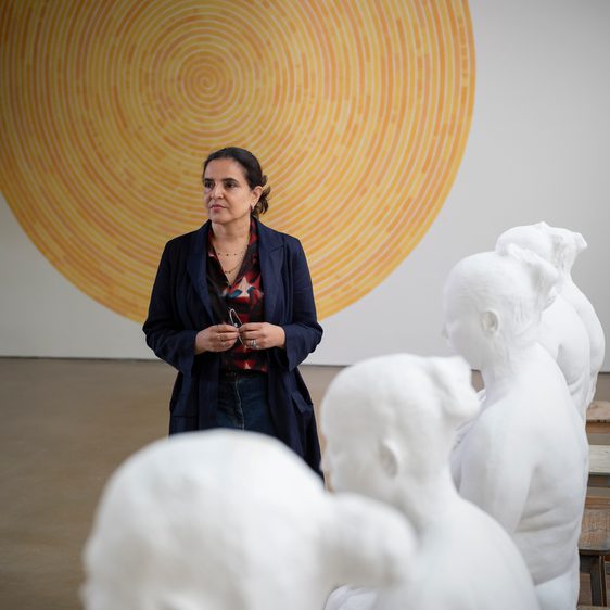 A woman wearing a black suit standing in front of a yellow spiral wall-based artwork. White plaster figures are in the foreground.